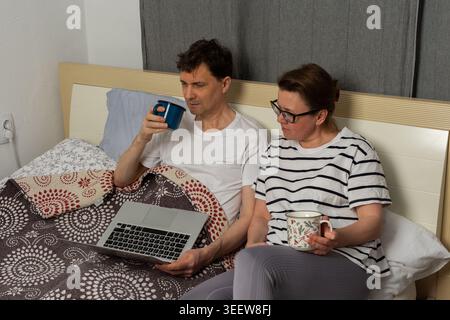 Un homme et une femme sont assis ensemble sur un lit. Ils tiennent tous les deux des boissons. L'homme regarde un ordinateur portable, tandis que la femme semble lire quelque chose sur son téléphone dans leur maison Banque D'Images
