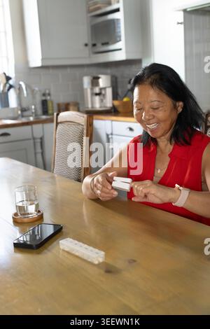 Femme asiatique senior assise dans la cuisine, boîte à pilules ouvrant le dessus rouge, téléphone et eau, espace de copie Banque D'Images