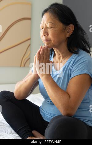 Femme asiatique senior méditant sur le lit à la maison portant un tee-shirt bleu clair et des leggings noirs Banque D'Images