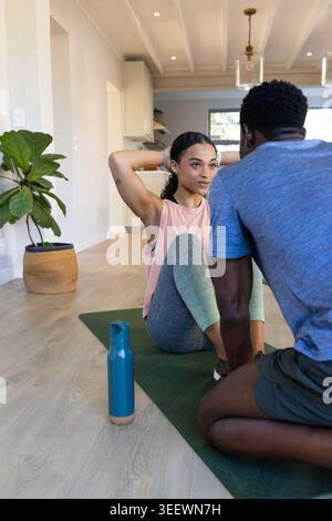 Couple africain exerçant dans des vêtements de sport à la maison, femme faisant sit-ups sur tapis vert, homme tenant les pieds Banque D'Images