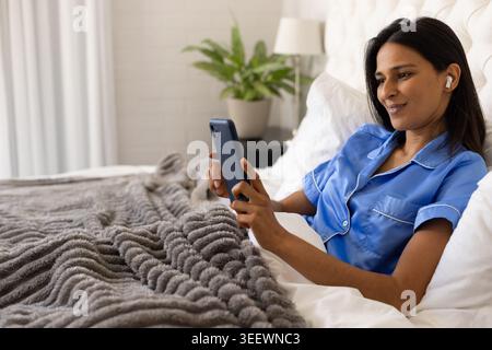 Femme indienne couchée dans la chambre portant le dessus en satin bleu tenant smartphone avec écouteur, espace copie Banque D'Images