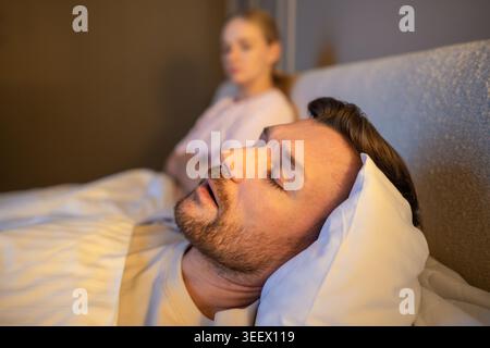 Homme dormant dans le lit et ronflant fort tandis que la femme frustrée assise à côté avec l'expression épuisée. Banque D'Images