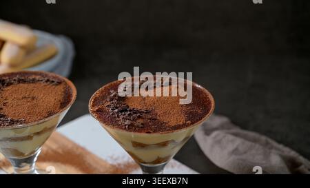 Des portions individuelles de tiramisu crémeux sont servies dans d'élégants verres à cocktail à tige sur un fond texturé foncé. Le dessert comporte visibl Banque D'Images