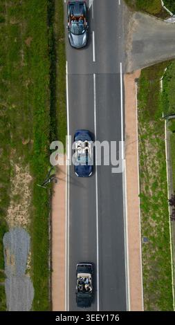 Vue aérienne de trois cabriolets avec des personnes à l’intérieur conduisant le long d’une route pavée bordée d’herbe verte et de végétation à la Chapelle-de-Guinchay, Banque D'Images