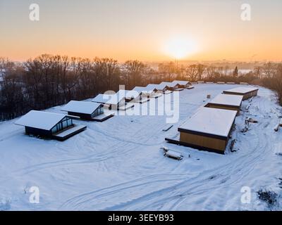 Rangée de cabines à ossature en bois modernes dans un paysage enneigé au coucher du soleil, vue aérienne du développement de la station éco avec des maisons minimalistes et une lumière chaude créant une atmosphère hivernale paisible Banque D'Images