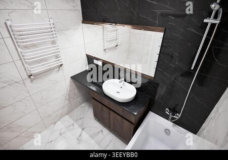 Vue panoramique sur la salle de bains moderne avec lavabo, baignoire et porte-serviettes chauffant. Salle de bain raffinée avec mur d'accent noir, carrelage aspect marbre, meuble-lavabo en bois et douche chromée Banque D'Images