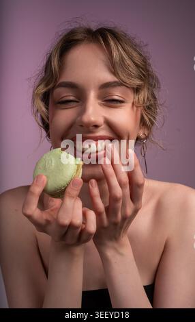 Femme souriante tenant un macaron vert mordu, savourant le goût sucré avec les yeux fermés sur un fond violet Banque D'Images