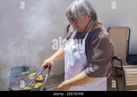 Femme dans un tablier cuisant des légumes et des saucisses sur un barbecue fumant, préparant un repas extérieur sain Banque D'Images