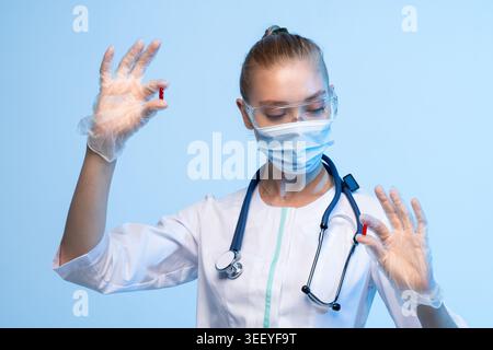 Femme médecin ou scientifique tenant deux capsules pharmaceutiques rouges pour examen, portant un équipement de protection individuelle Banque D'Images