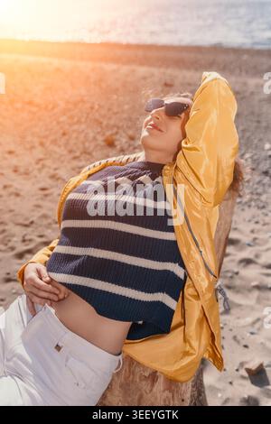 Femme plage détente femme heureuse portant imperméable jaune et lunettes de soleil se détend sur la plage de sable sous le soleil doré chaud Banque D'Images