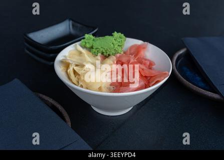 Gingembre rose et blanc mariné et bol de wasabi sur table sombre, condiments japonais traditionnels servis avec sushi, sauces empilées et serviettes sur bl Banque D'Images