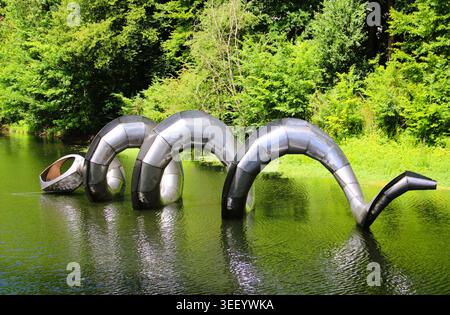 Serpent métallique serpente à travers l'eau verte tranquille. Une forêt luxuriante encadre la surface réfléchissante de la sculpture. A peine visible, la tête du serpent pairs f Banque D'Images