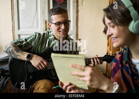 Jeune homme caucasien adulte jouant de la guitare électrique tandis que jeune femme adulte portant des écouteurs prenant des notes, collaborant à l'écriture de chansons. Idéal pour l'éducation musicale, le travail d'équipe créatif Banque D'Images