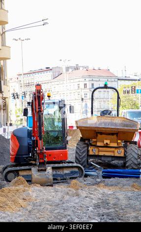 Excavatrice compacte et mini tombereau travaillant sur un site de réparation de routes urbaines avec du sable, des tuyaux et des bâtiments historiques en arrière-plan. Banque D'Images