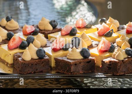 Assortiment de brownies au chocolat et de bouchées crémeuses de cheesecake à la mangue garnies de fraises fraîches, de bleuets et de crème fouettée à la lumière naturelle chaude. Banque D'Images