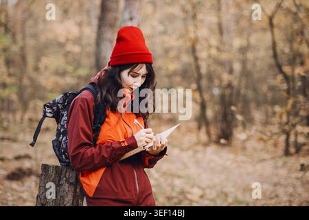 Femme se tient dans la forêt d'automne et écrit dans un cahier tout en s'appuyant sur la souche d'arbre. Travailler de n'importe où, espace de travail extérieur indépendant, nomade Banque D'Images