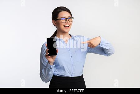 Woman in Glasses pointe vers smartphone avec excitation dans un cadre minimaliste Banque D'Images