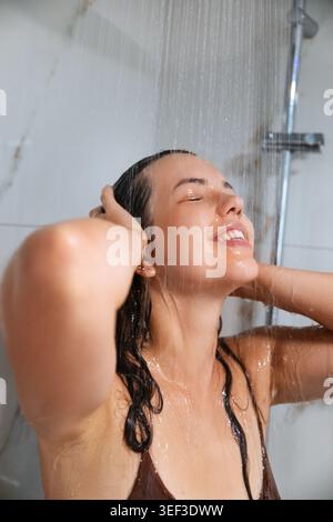 Jeune femme se relaxant sous la douche avec une apparence fraîche radieuse. Femme attrayante nettoyant les longs cheveux mouillés en utilisant la routine quotidienne de soin de la peau. Corps de bien-être Banque D'Images