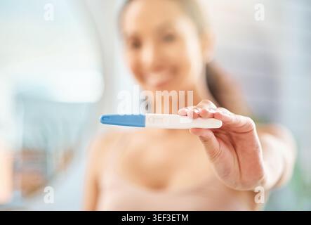 Femme heureuse, main et test de grossesse à la maison pour l'attente, la maternité ou de bonnes nouvelles dans la salle de bain. Personne féminine, sourire ou bâton de maternité avec examen Banque D'Images