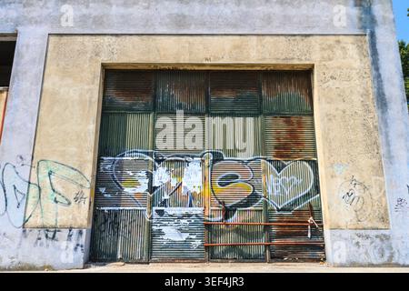 Une vieille porte d'entrepôt en métal ondulé altéré recouverte de graffitis décolorés et d'affiches déchirées, placée dans une façade de bâtiment en béton. Cet ima texturé Banque D'Images
