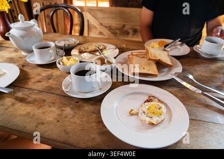 Petit déjeuner traditionnel sri-lankais servi sur une table rustique en bois, avec oeuf frit, pain grillé, beurre, confiture, et des tasses de thé Banque D'Images