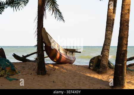 Bateau de pêche traditionnel en saillie reposant sur la plage de sable sous les palmiers avec les vagues de l'océan à Tangalle, Province du Sud, Sri Lanka Banque D'Images