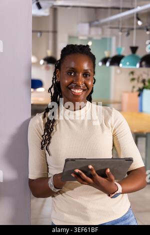 Femme afro-américaine appuyée contre la colonne dans le bureau, souriante et tenant la tablette avec smartwatch Banque D'Images