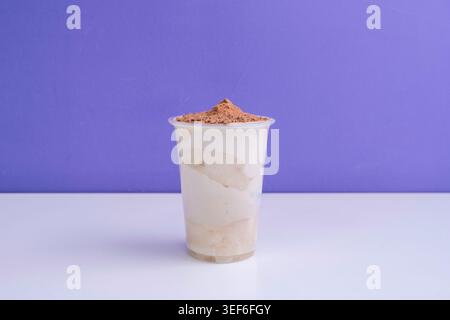 Une boisson blanche crémeuse surmontée de crumbles bruns remplit une tasse en plastique sur une surface blanche sur un fond violet. Banque D'Images