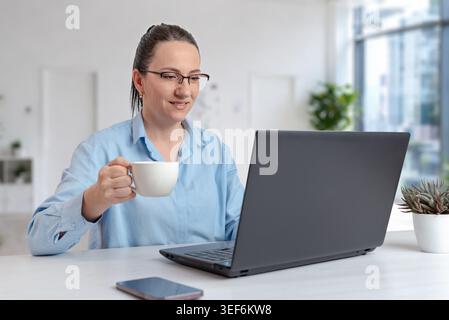 Femme d'affaires buvant du café tout en travaillant sur ordinateur portable dans un espace de travail de bureau lumineux, représentant un style de vie équilibré de travail à distance et une productivité moderne Banque D'Images