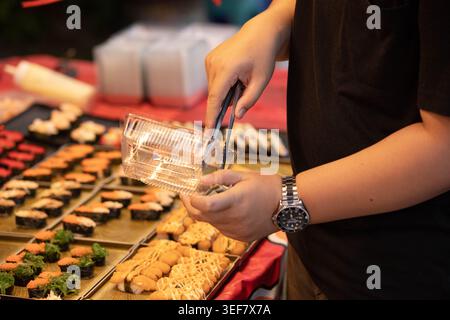 Homme utilisant des pinces pour ramasser des sushis dans une boîte en plastique, visiteurs naviguant sur le marché touristique populaire, gros plan. Banque D'Images