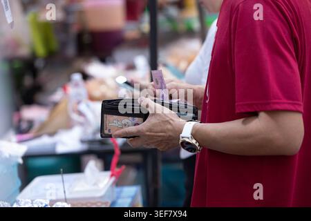 PHAYAO, THAÏLANDE-APR 11, 2026 : homme payant pour des marchandises avec des billets de banque, gens faisant des achats dans le marché en plein air, gros plan, Chiang Kham. Banque D'Images