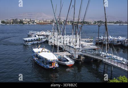 Louxor, Égypte - 27.07.2025. Les bateaux à voile traditionnels felouques et autres bateaux d'excursion sont amarrés à une jetée animée sur le Nil, prêts pour les touristes Banque D'Images