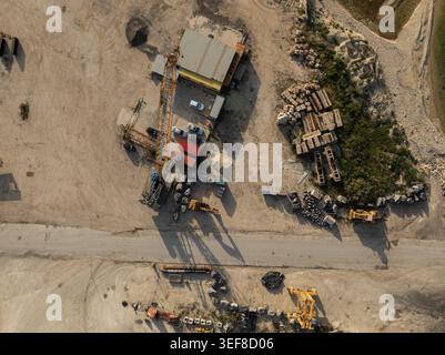 Vue aérienne d'un chantier industriel avec une grande grue jaune, de la machinerie lourde et des véhicules stationnés sur un sol sablonneux à Priolo Gargallo Banque D'Images