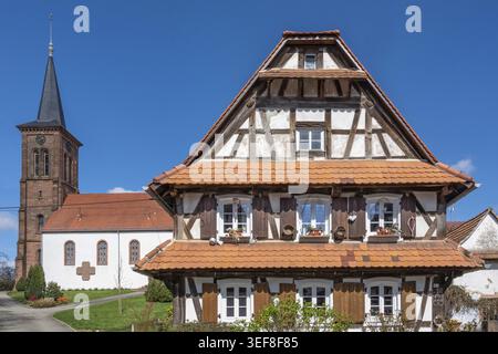 Une grande maison traditionnelle à colombages en plâtre blanc avec poutres sombres, toits de tuiles rouges et jardinières se dresse devant la tour de l'église du P. Banque D'Images