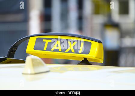 Gros plan d'un panneau de taxi jaune et noir classique monté sur le toit d'une voiture de couleur crème. Le mot « TAXI » est clairement visible face à un urba flou Banque D'Images