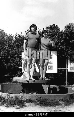 Sloviansk, 1983 ans, camp pionnier de Vishnegorsk à Slavkurort. Deux conseillers du camp se tiennent fièrement sur le dernier étage d'une fontaine en pierre. L'homme est pieds nus, tandis que la femme à côté de lui tient un ballon de football. Les deux portent des t-shirts et des shorts d'athlétisme foncés assortis. Le ciel lumineux d'été et le feuillage du parc environnant encadrent ce portrait imposant. La photographie reflète la forme physique, le travail d'équipe et le style de vie énergique du personnel des jeunes pendant les vacances d'été dans le Donbass paisible. Banque D'Images