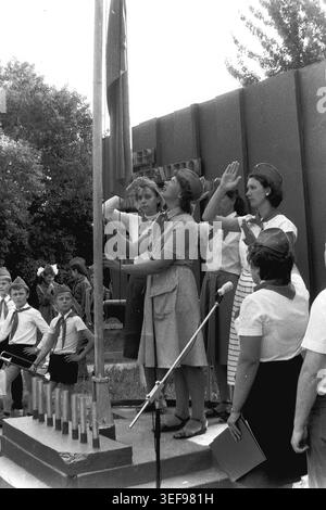 Sloviansk, 1983 ans, camp pionnier de Vishnegorsk situé à Slavkurort. La cérémonie d'ouverture officielle du camp d'été comprend un rituel solennel de lever de drapeau. Les leaders féminins de la jeunesse, portant d'élégantes robes d'été avec des casquettes et des cravates pionnières, participent à hisser le drapeau. Un conseiller tire la corde tandis que les autres rendent des salutations respectueuses des pionniers. Cette image historique évocatrice capture la discipline idéologique, les traditions formelles et l'environnement éducatif structuré des organisations de jeunesse soviétiques dans le Donbass pacifique. Banque D'Images