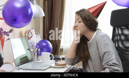 Triste femme célibataire célébrant son anniversaire par la fête virtuelle d'appel vidéo. Assis seul avec un ordinateur portable et gâteau avec une bougie. Lieu de travail authentique. Coron Banque D'Images