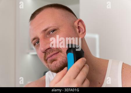Beau jeune homme se rasant le visage avec un rasoir électrique dans le miroir de salle de bain pendant la routine du matin. Toilettage masculin, soin de la peau et concept de soins personnels. Banque D'Images