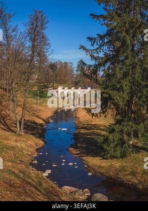 La lumière du soleil jaillit de l'eau calme de la rivière. Les arbres sont hauts sur les deux rives. Le pont vole gracieusement à travers le ruisseau. Le ciel bleu s'étend au-dessus de la scène. Les feuilles d'automne couvrent le sol. Oranienbaum Banque D'Images