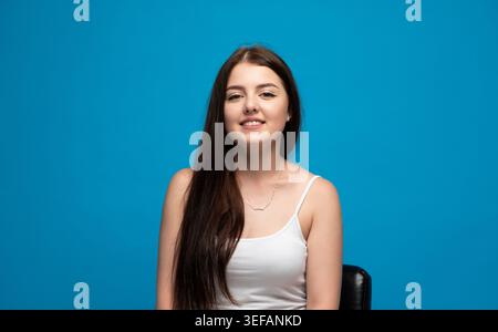 Jeune femme souriante, regardant la caméra avec de longs cheveux bruns sur fond bleu, se sentant heureuse et positive Banque D'Images