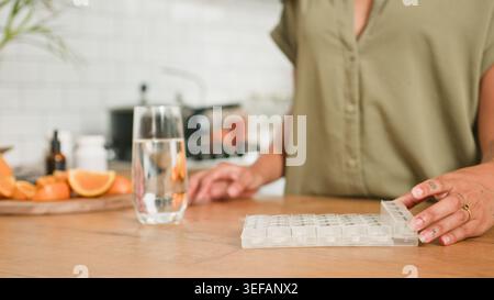 Femme utilisant l'organisateur hebdomadaire de pilules sur le comptoir de cuisine avec verre d'eau et d'agrumes Banque D'Images