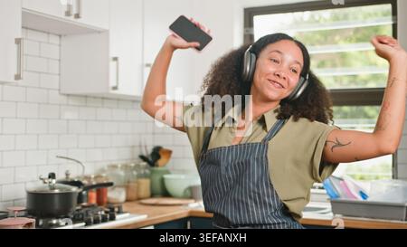 Femme heureuse dansant dans la cuisine portant casque et tablier tout en tenant smartphone Banque D'Images