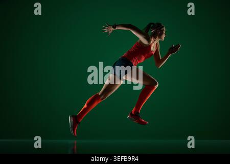 Coureuse féminine concentrée sprintant vers l'avant sur le sol vert foncé du studio. Banque D'Images