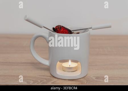 Mug à fondue en céramique blanche à la fraise trempée dans du chocolat, réchauffée par une bougie allumée, reposant sur une surface en bois clair Banque D'Images