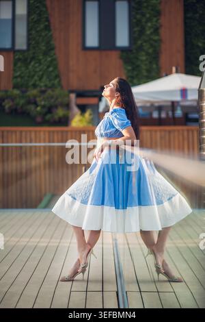 Femme élégante avec de longs cheveux en tenue bleue et blanche se détend sur la terrasse ensoleillée. Elle s'appuie sur une balustrade en verre, face au soleil, avec son reflet visible. Banque D'Images