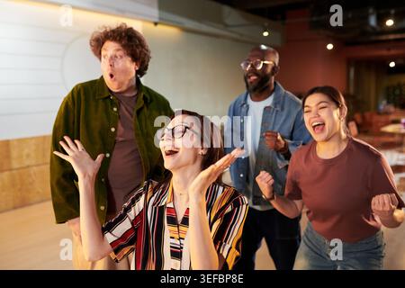 Divers jeunes adultes acclamant tout en regardant la présentation au bureau, célébrant le succès et la réussite de l'équipe. Idéal pour le travail d'équipe en entreprise, la culture de démarrage, le marketing des objectifs d'entreprise Banque D'Images