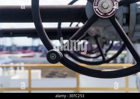 Grande roue de valve noire sur tuyau industriel. L'arrière-plan montre une installation industrielle floue. La roue a des marques rouges et blanches. L'image montre un gros plan de HE Banque D'Images