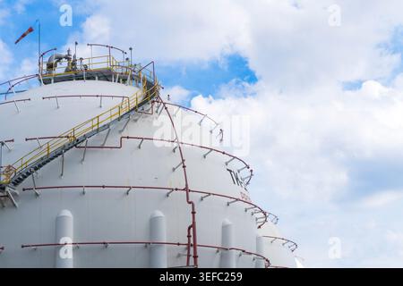 Grand réservoir de stockage blanc sous ciel bleu avec des nuages. Les canalisations rouges et les rampes de sécurité jaunes enveloppent la surface incurvée du réservoir. Des passerelles métalliques mènent au toit Banque D'Images