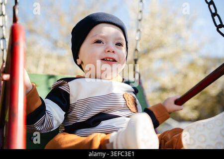 Bébé garçon caucasien assis sur l'oscillation de terrain de jeu dans le parc résidentiel ensoleillé. Enfance et mode de vie en plein air. Banque D'Images